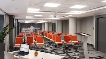 Une salle de conférence avec des rangées de chaises orange, une table blanche sur laquelle se trouve un ordinateur portable et un pupitre.