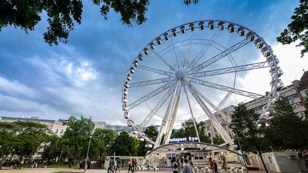Una noria grande en un parque público con un edificio que dice "Budapest Eye" al fondo.