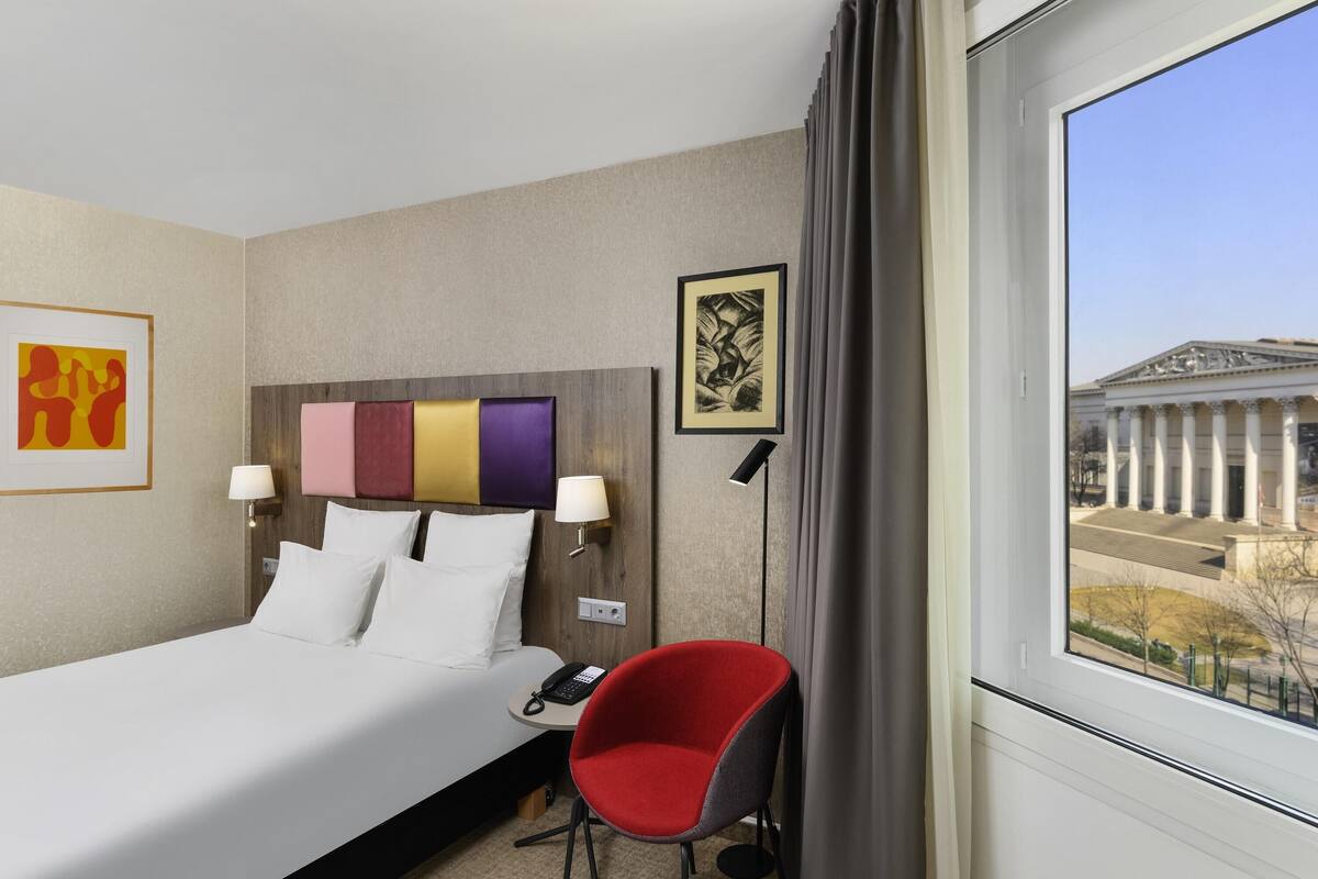 A hotel room with a bed, a red chair, a lamp, and a view of a building with columns.