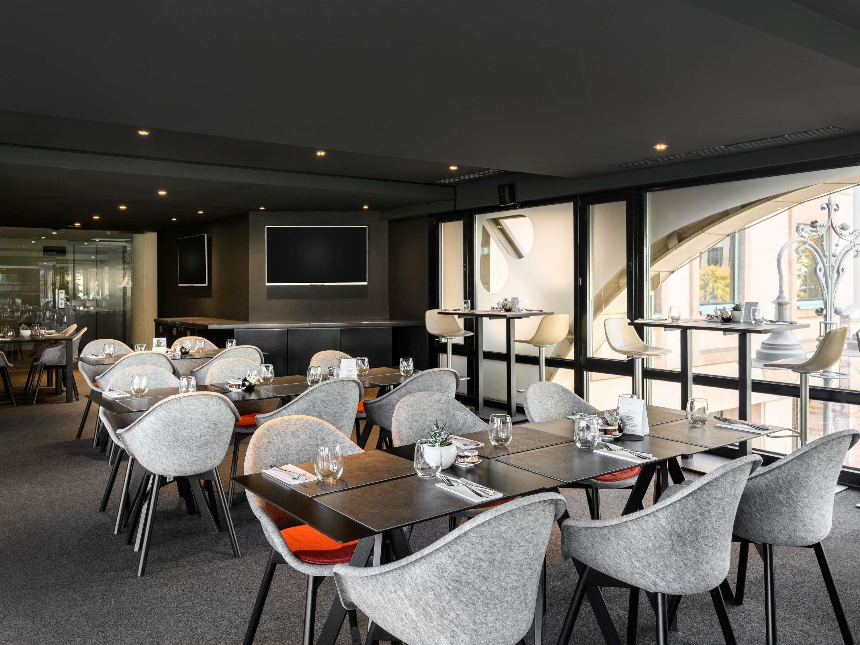 A modern dining area with grey upholstered chairs, dark tables, and a large window offering a view of the outdoors.