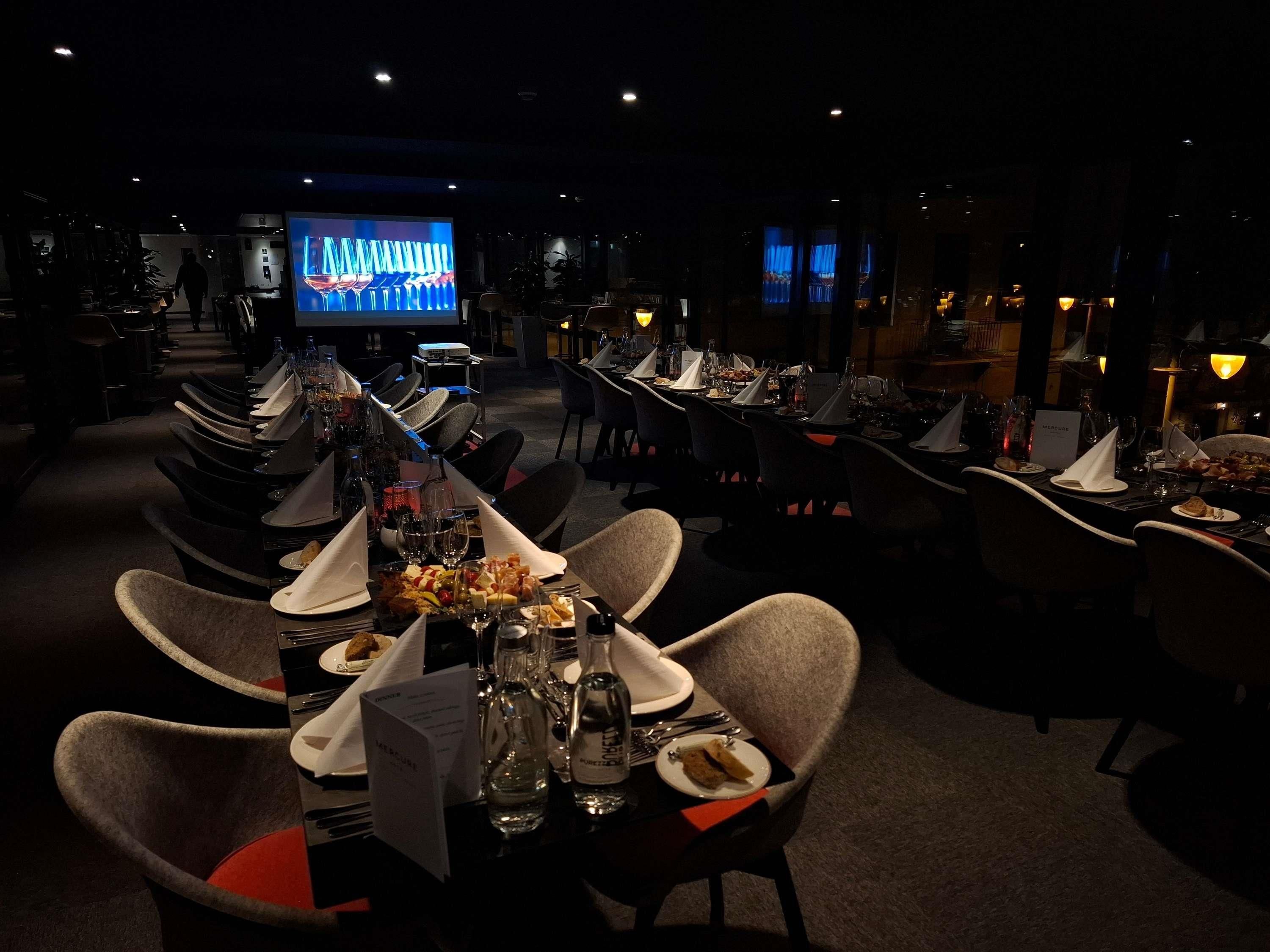 A dimly lit restaurant with tables set for dining, a television screen in the background, and a dark ambiance.