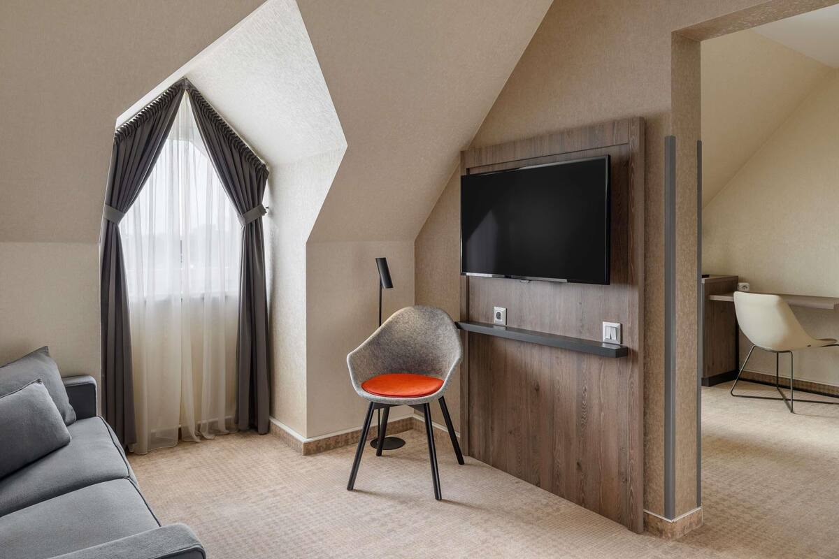 A modern hotel room with a large TV, a grey sofa, a chair with an orange cushion, and a wooden accent wall.