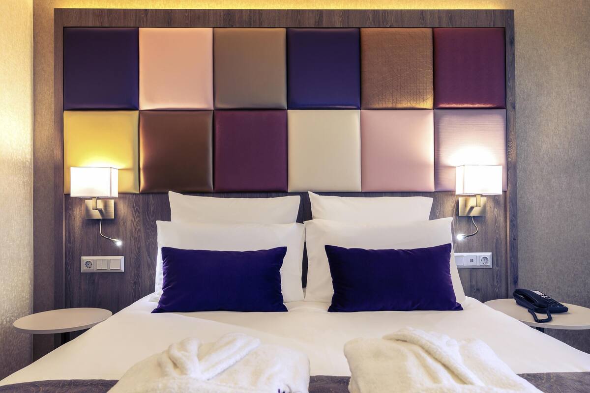 A hotel room with a large bed, two bedside tables, a headboard with multiple panels, and a wall socket.