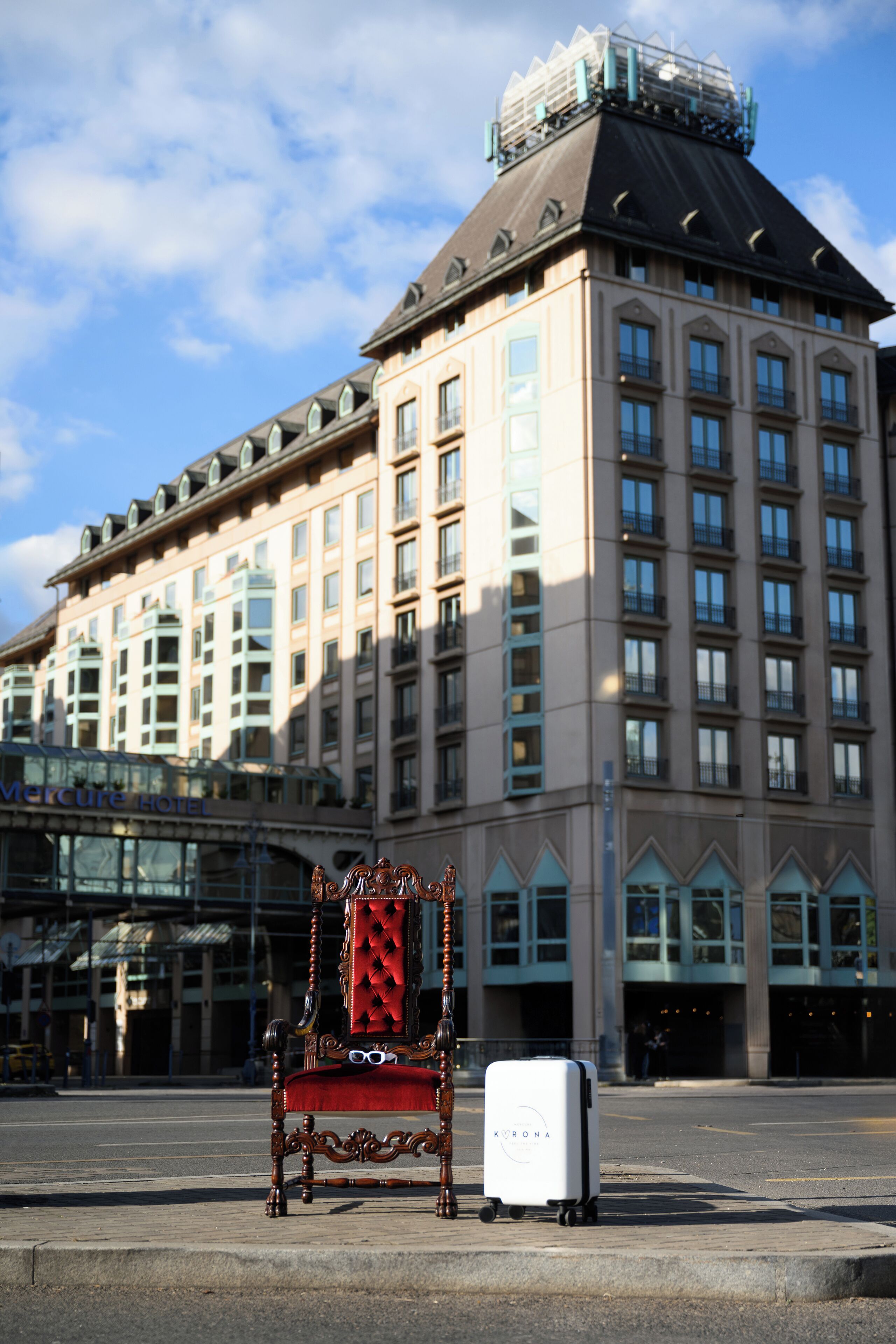 A large hotel building with a classic architectural style, featuring multiple windows and balconies. In front of the building, there is a vintage-style chair and a suitcase on the sidewalk.