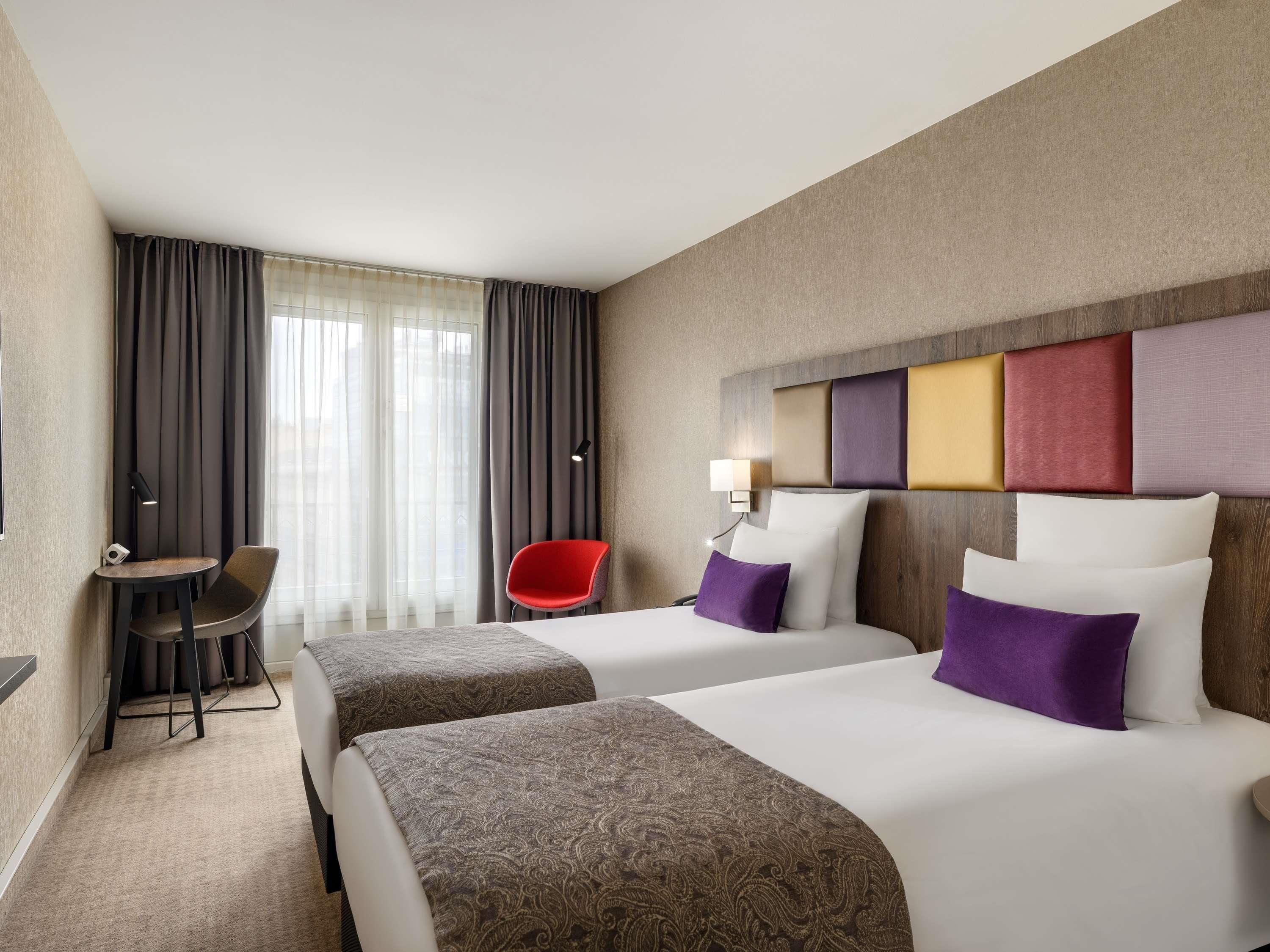 Een hotelkamer met twee bedden, een bureau, een stoel en een groot raam met gordijnen.