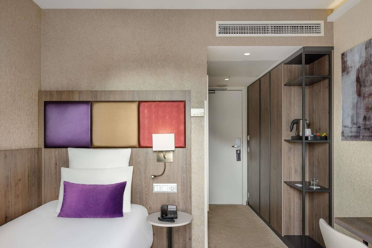A hotel room with a bed, bedside table, lamp, and a view of the hallway.