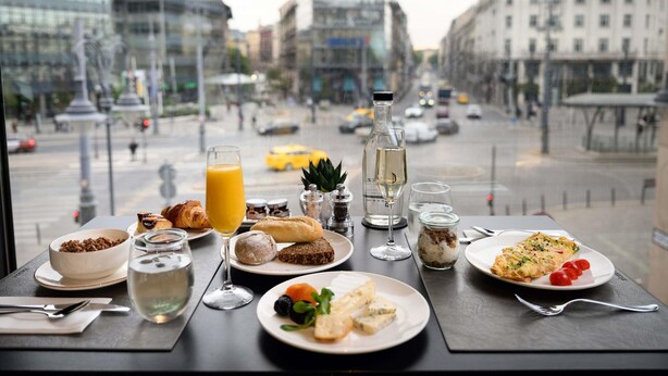 Uma mesa com itens de café da manhã e bebidas, com vista para uma rua urbana com edifícios e veículos.