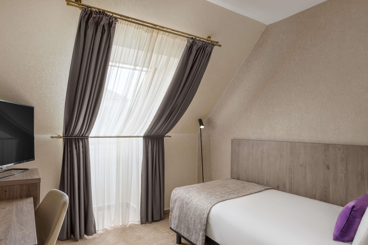 Una habitación de hotel con cama, escritorio con televisor, una silla y ventana con cortinas.