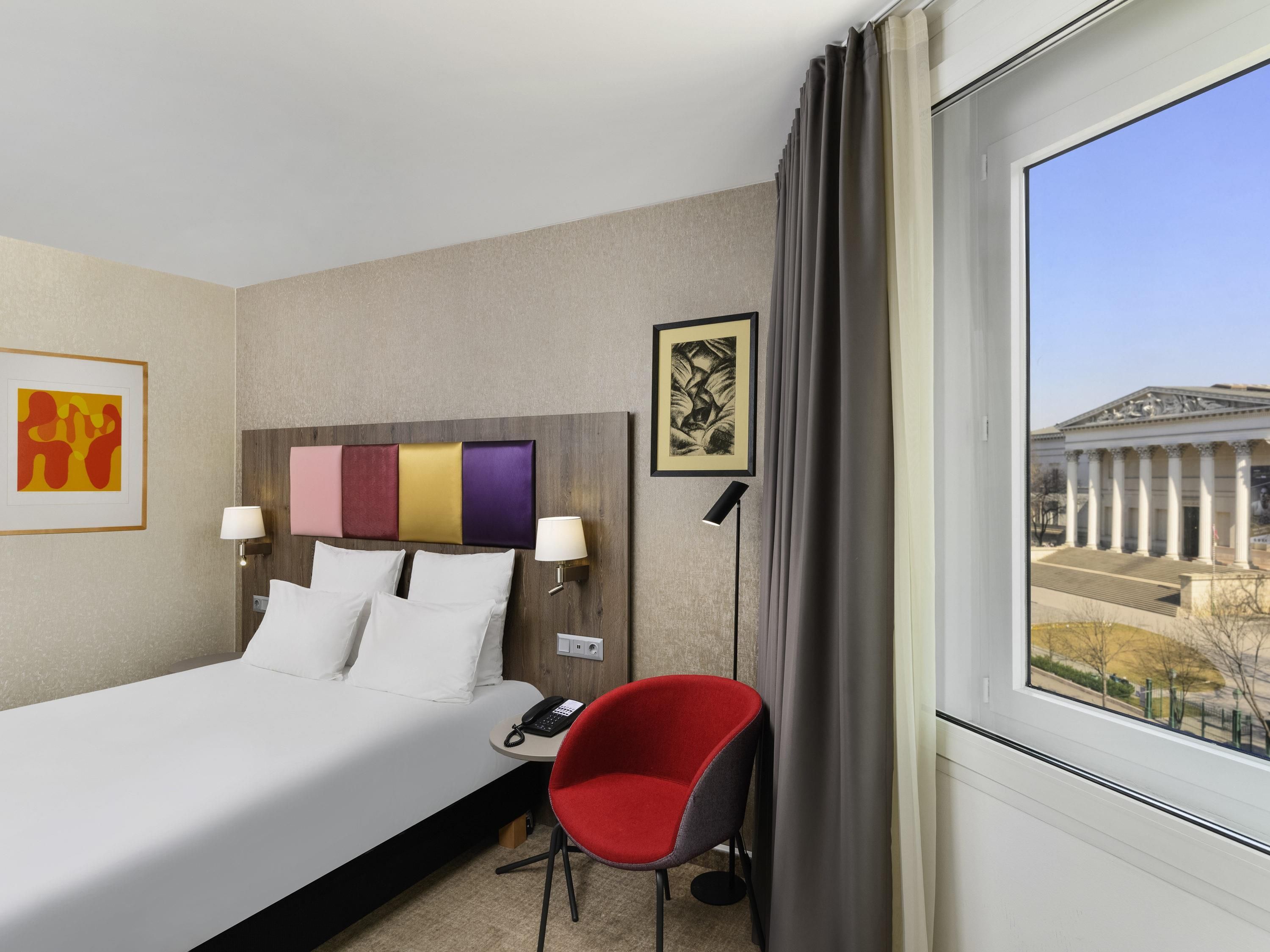 Een hotelkamer met een bed, een rode stoel, een lamp en uitzicht op een gebouw met kolommen.