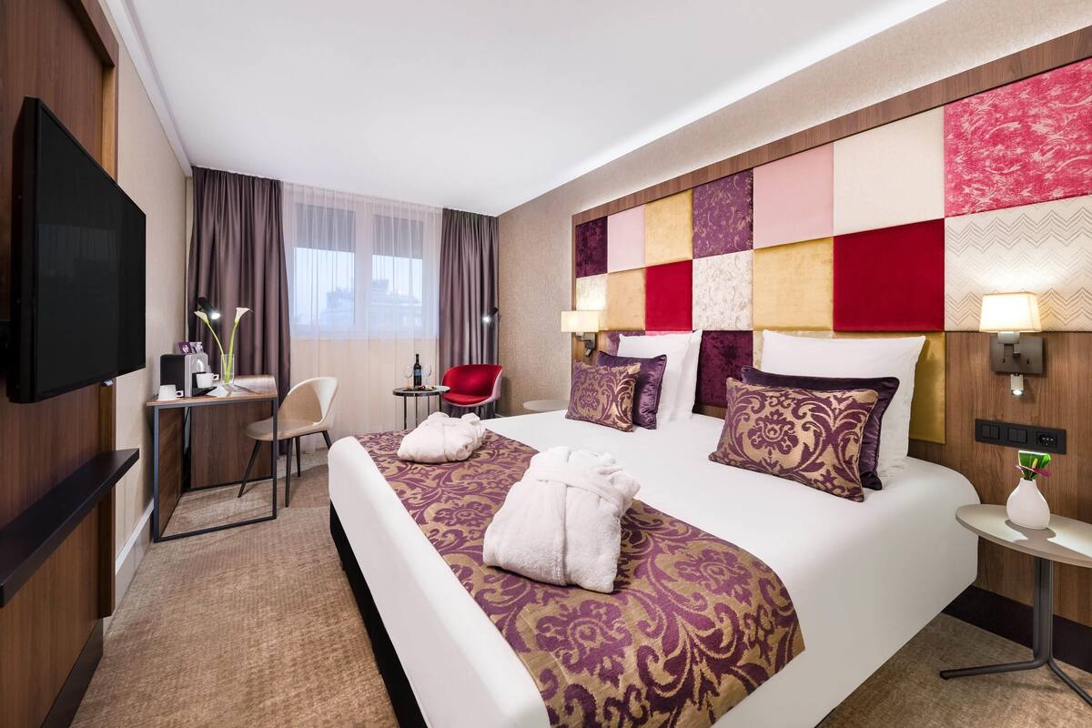 A hotel room with a large bed, a desk, a chair, a TV, and a window with curtains.
