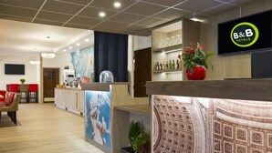 Un espace d’accueil d’hôtel au design moderne, comprenant un comptoir de réception, un écran de télévision affichant le nom de l’hôtel, un réfrigérateur à vin fixé au mur et un coin salon avec des chaises rouges.