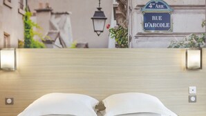 Une chambre d’hôtel avec une tête de lit en bois, un lit avec des oreillers blancs, un interrupteur près du lit et une pancarte indiquant « 4e arrondissement, rue d’Arcole ».