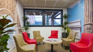 Un hall d’entrée avec des fauteuils beiges et rouges disposés autour d’une table centrale, de grandes fenêtres et une fresque représentant un bâtiment.