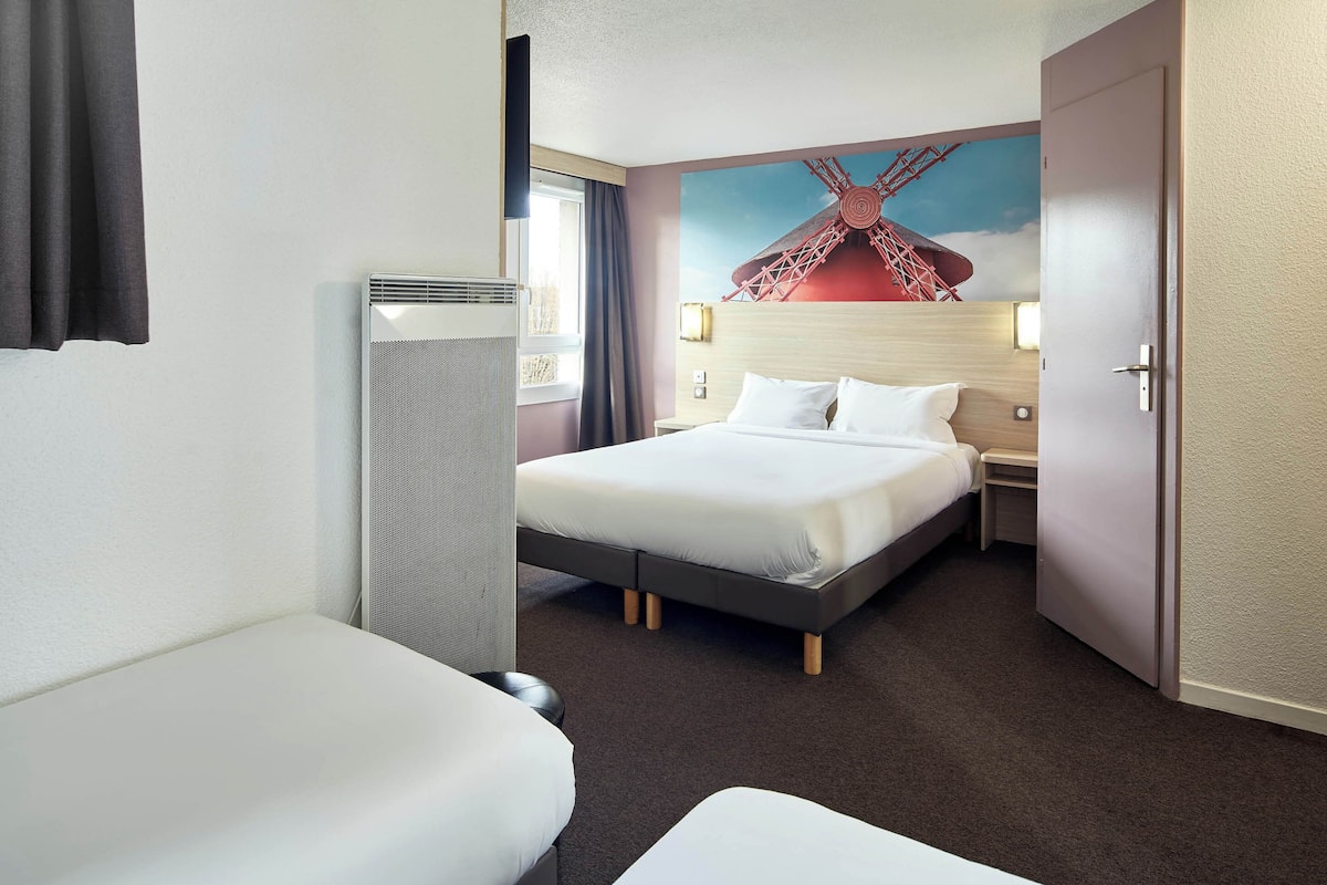 A hotel room with a bed, a bedside table, a lamp, a window with curtains, and a wall painting of a windmill.
