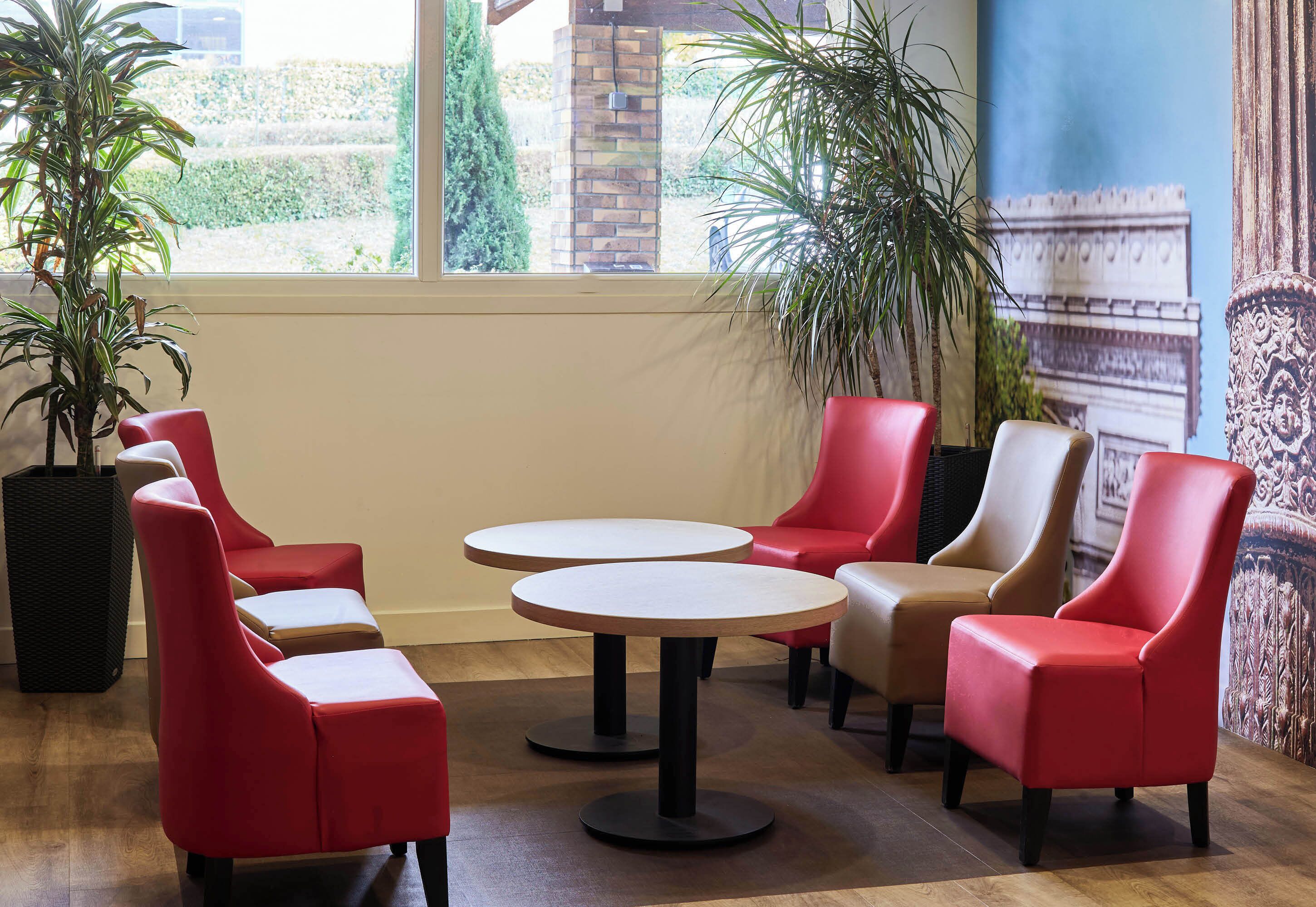 A seating area with red and beige chairs around two round tables, large potted plants, and a mural of the Arc de Triomphe.