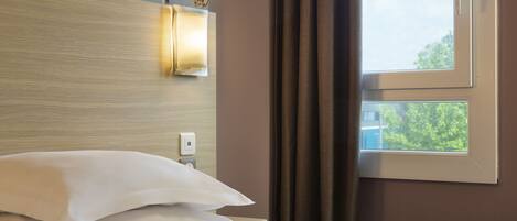 A hotel room with a bed, a nightstand, a wall-mounted light, a window with curtains, and a view of greenery outside.