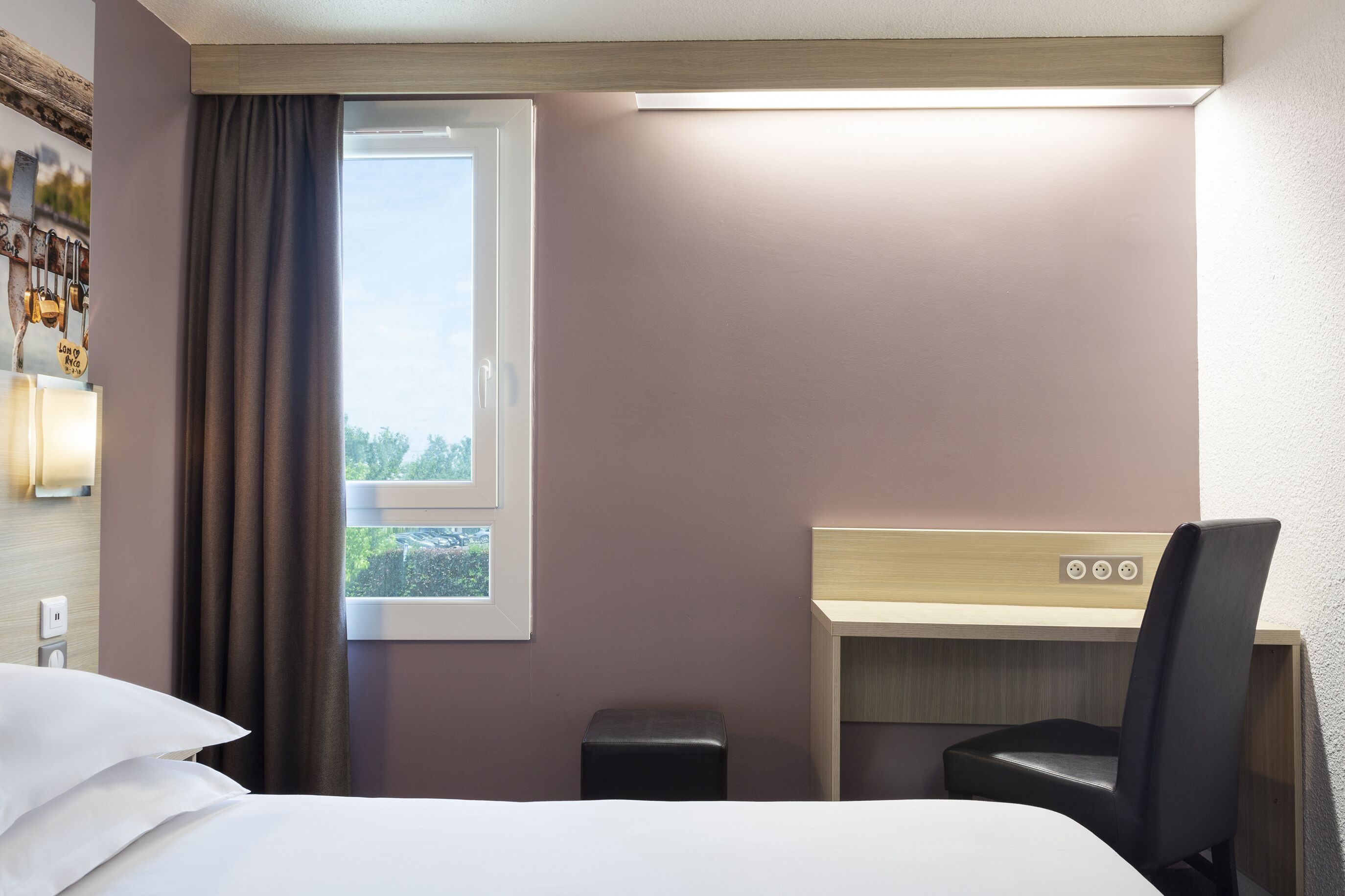 Habitación de hotel con cama, escritorio, silla, taburete pequeño, ventana con cortinas y lámpara fijada a la pared.