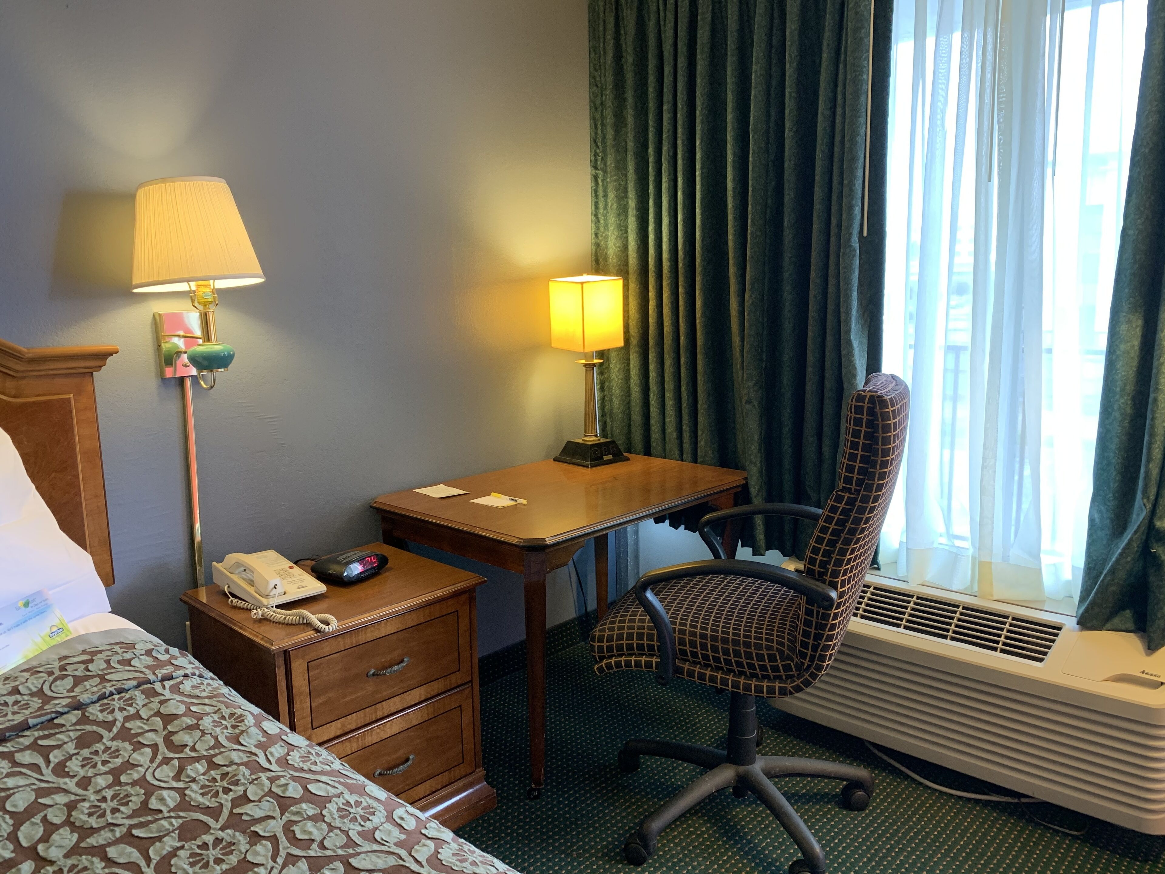 Habitación de hotel con cama, escritorio, silla, teléfono, lámpara y aire acondicionado.