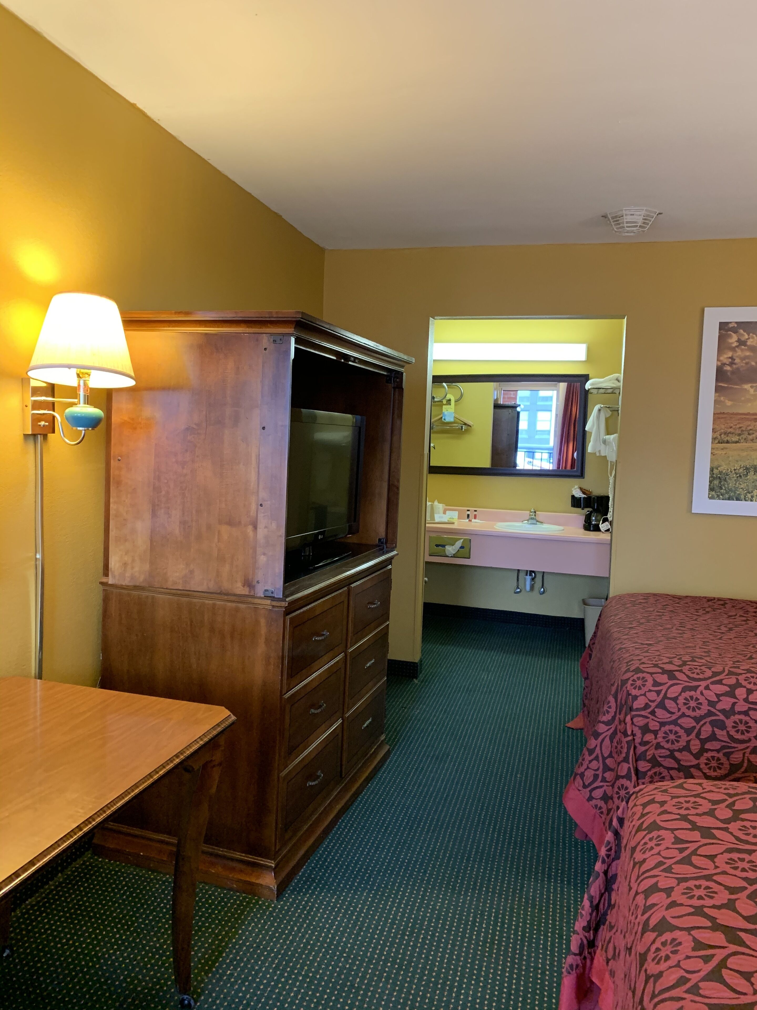 A hotel room with a wooden dresser, a television, a desk, a bed with a patterned bedspread, a bathroom with a sink and mirror, and a wall-mounted light fixture.