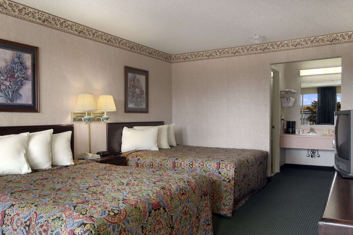 A hotel room with two beds, a desk with a television, a bathroom with a sink and mirror, and a floral-patterned bedspread.