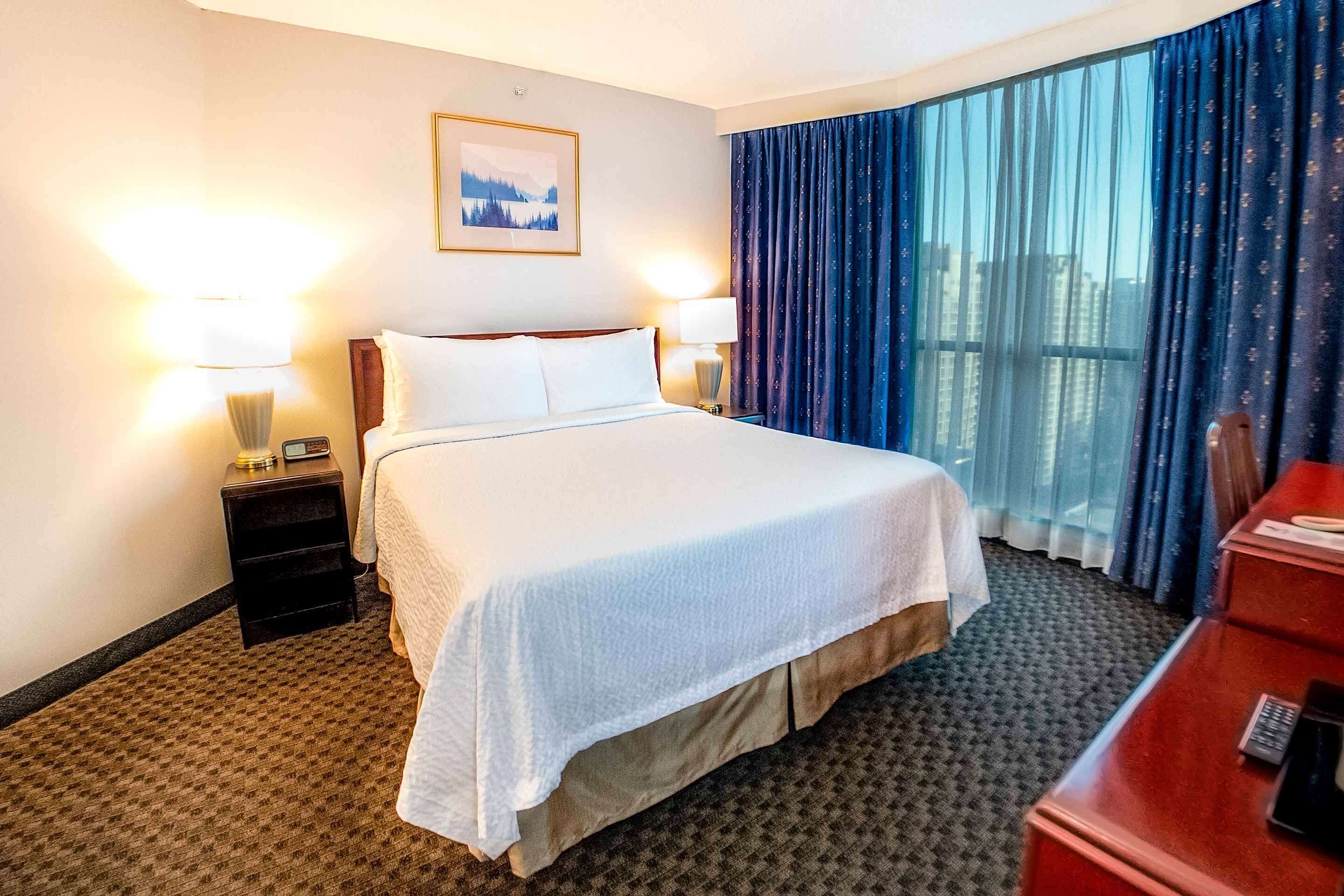A hotel room with a large bed, two bedside lamps, a nightstand, a desk with a phone, and a window with curtains.