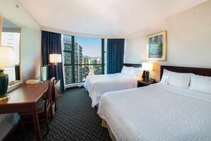 A hotel room with two beds, a desk, a chair, a lamp, and a large window offering a city view.