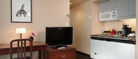 A compact hotel room with a wooden desk, a television, a small kitchenette with a microwave, and a framed artwork on the wall.