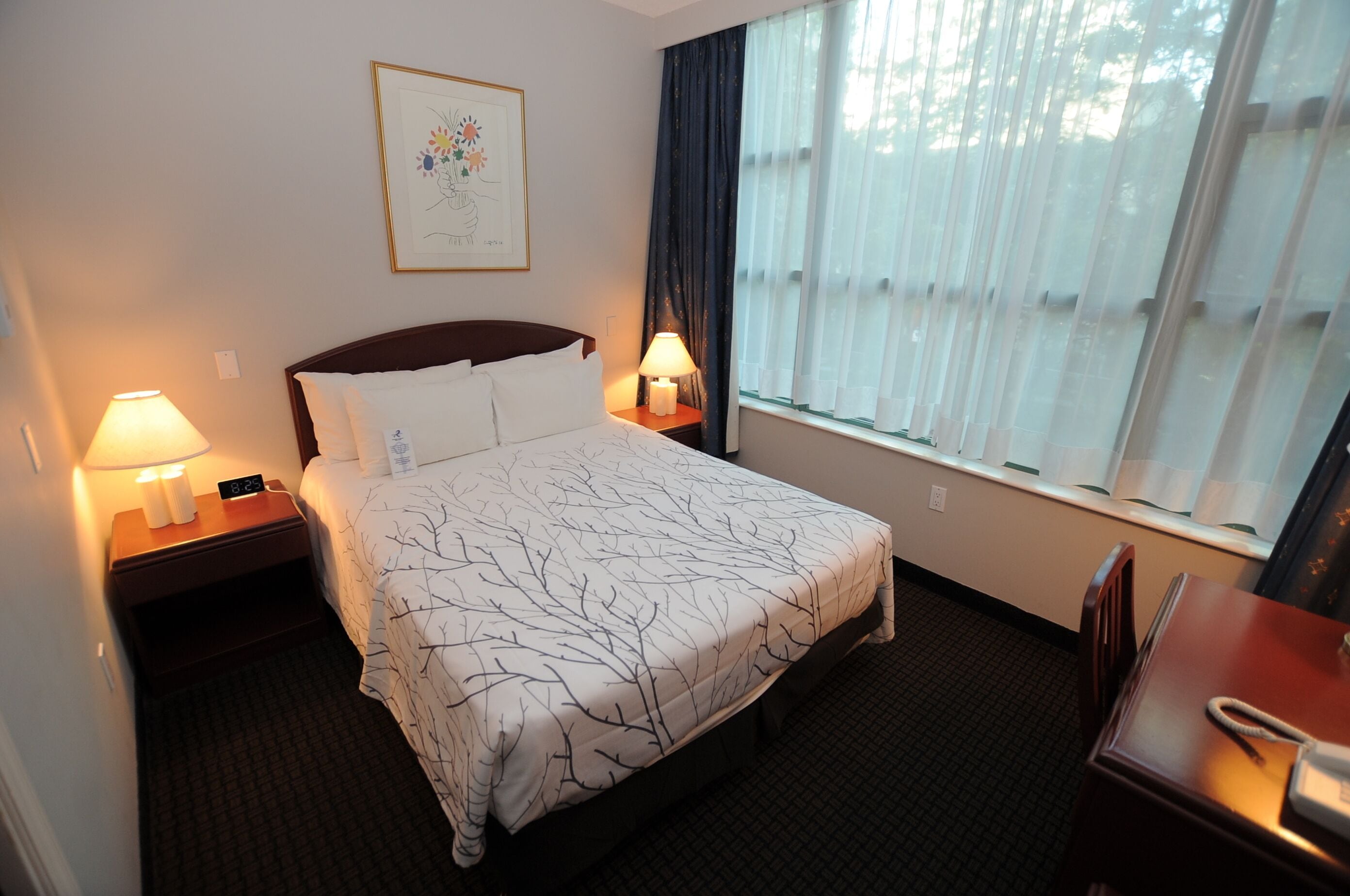 A hotel room with a bed, bedside lamps, a window with curtains, a desk, and a chair.