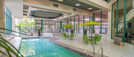 An indoor swimming pool with clear glass walls, green chairs, and umbrellas.