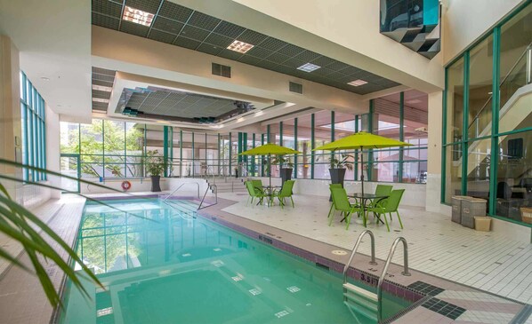 An indoor swimming pool with clear glass walls, green chairs, and umbrellas.
