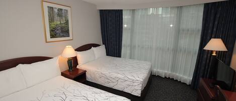 A hotel room with two beds, a nightstand with a lamp, a desk with a lamp, a framed picture on the wall, and a window with curtains.