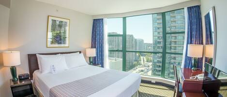 A hotel room with a large bed, two bedside tables, a desk with a chair, a large window with a view of high-rise buildings, and a framed picture on the wall.