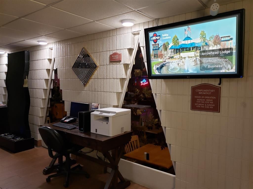 A hotel reception area with a computer monitor displaying the Arrowhead Lodge, a complimentary breakfast sign, and a business center sign.