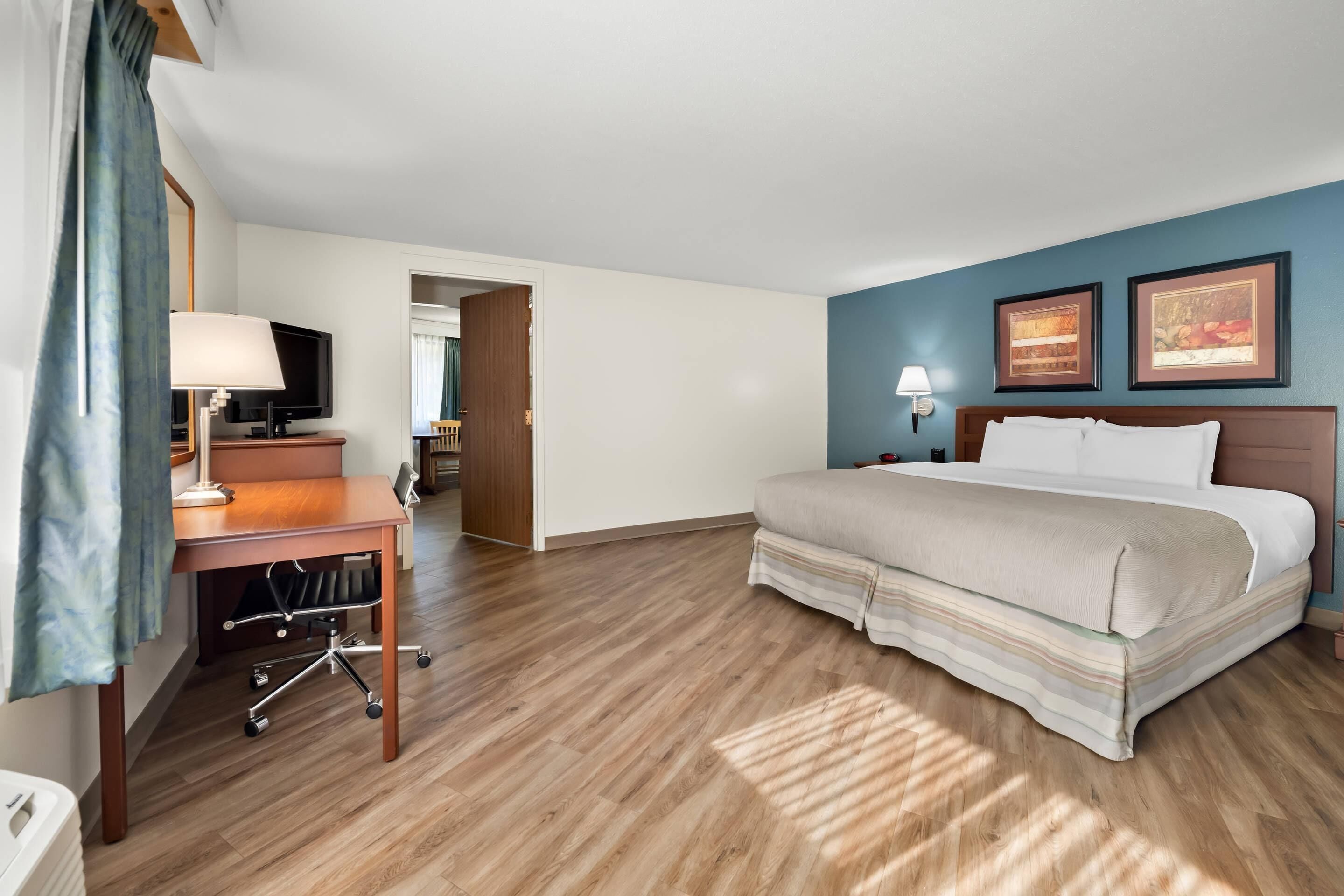 A hotel room with a bed, desk, chair, TV, and artwork on the wall.
