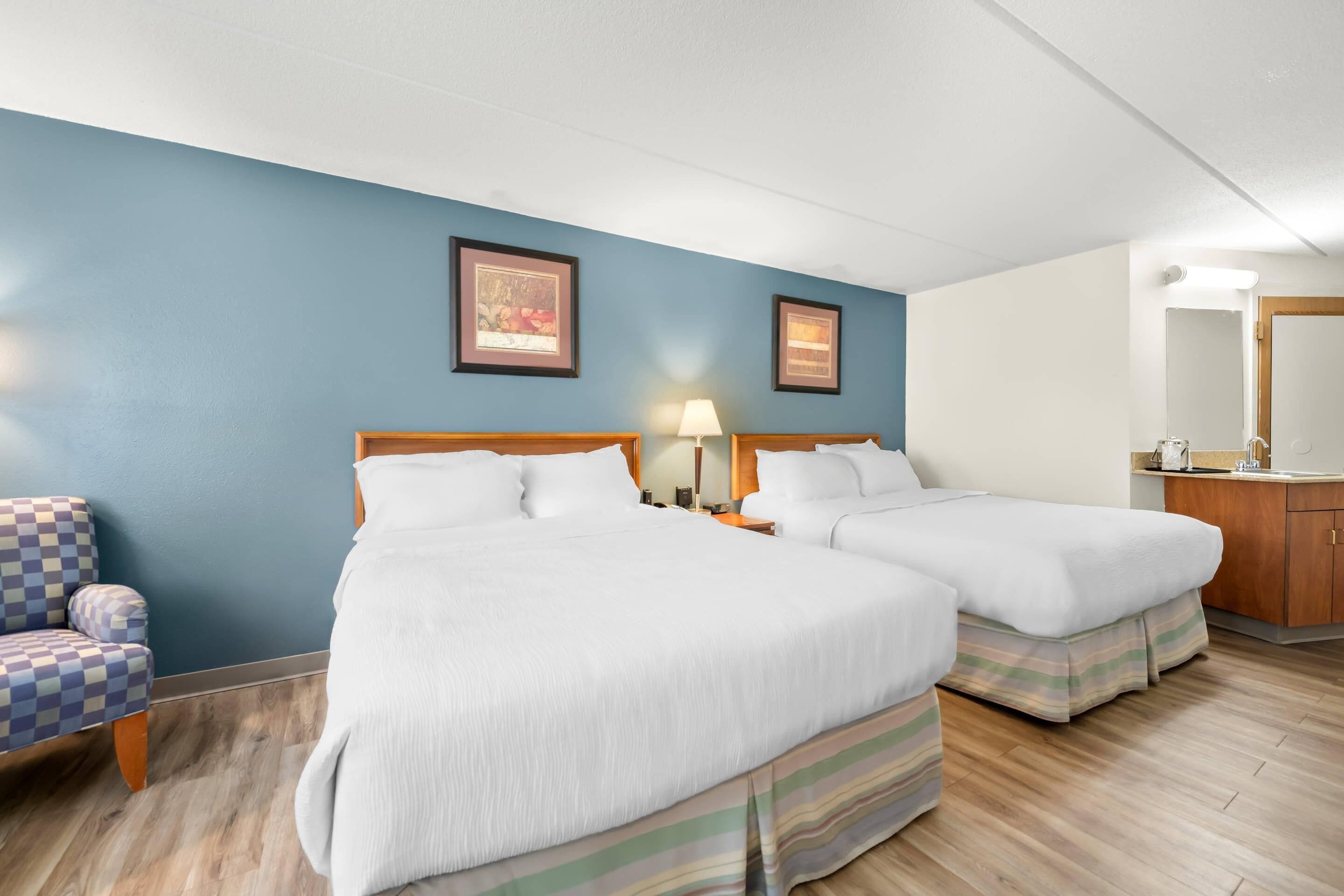 Habitación de hotel con dos camas, un sillón a cuadros, una mesita de noche con lámpara y un espejo.