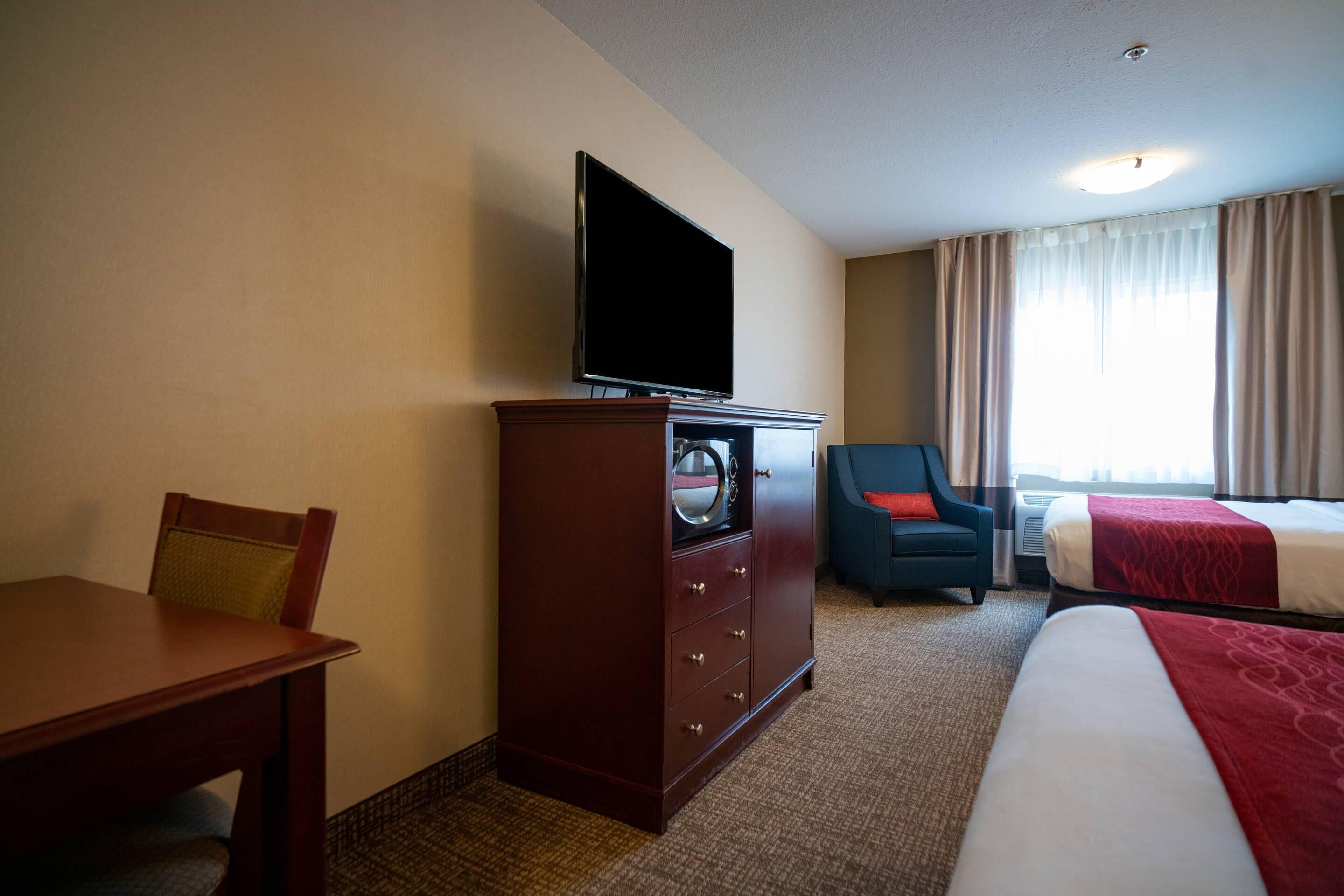 Habitación de hotel con televisor sobre un tocador, un escritorio, una silla, dos camas y una ventana con cortinas.