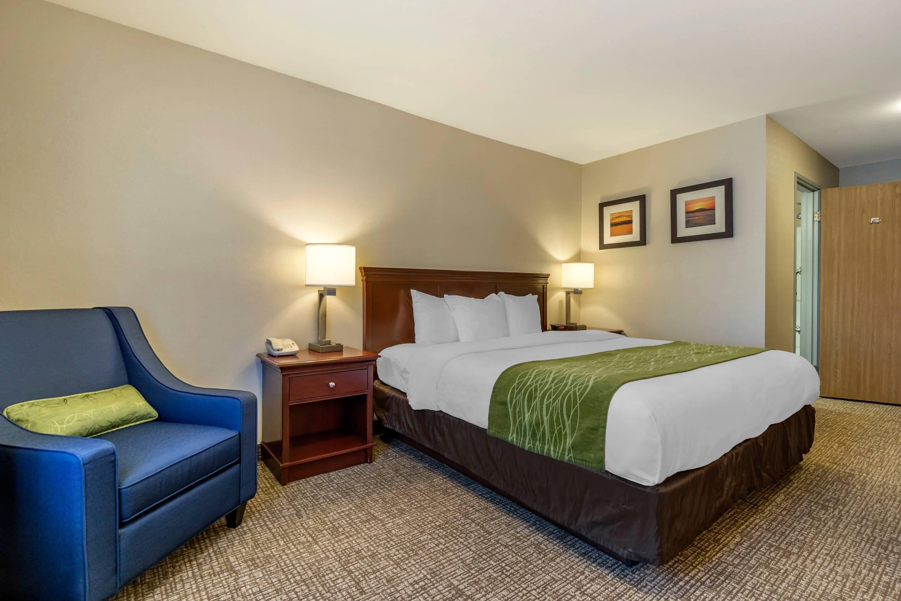 A hotel room with a large bed, a blue armchair, a nightstand, two wall-mounted lamps, and two framed pictures on the wall.