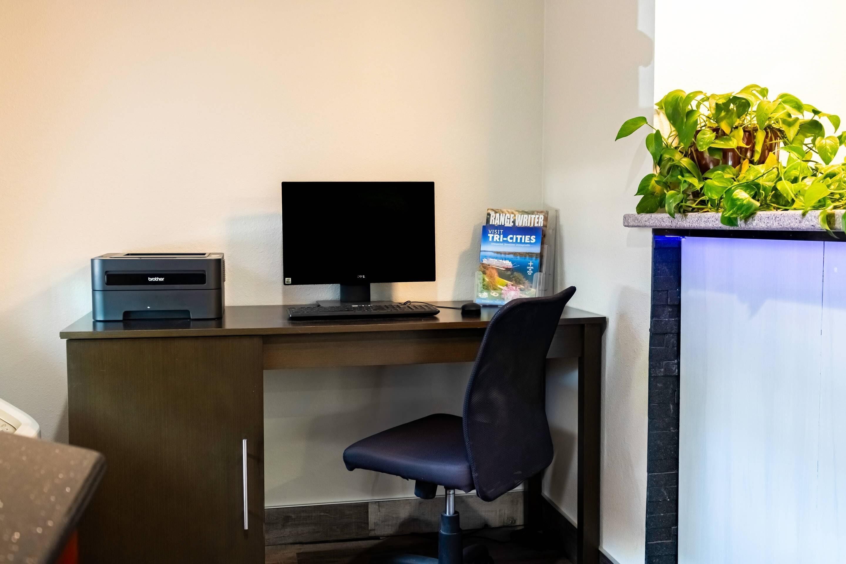 Un escritorio con una impresora, un monitor de computadora y una silla. Hay una planta en la pared y un libro titulado 