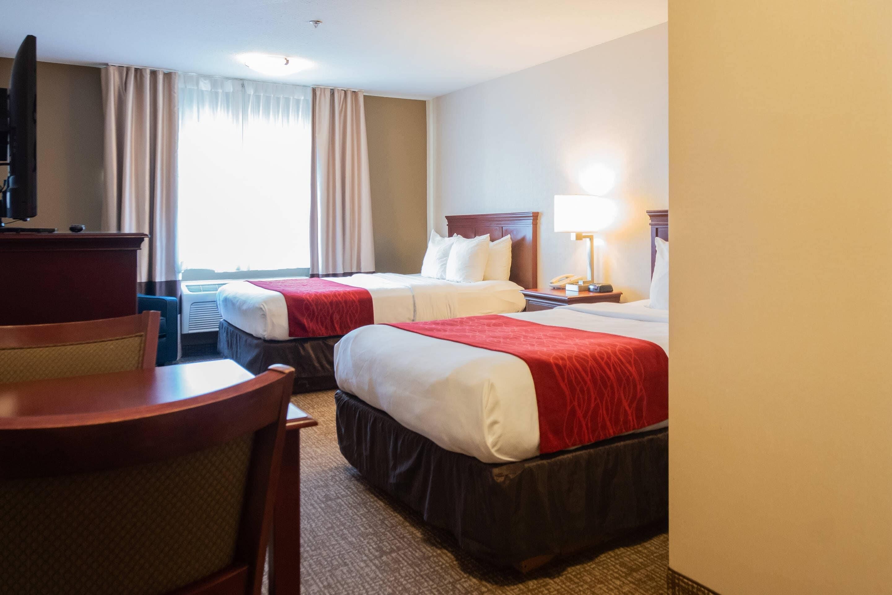A hotel room with two beds, a desk, a chair, a TV, and a lamp.