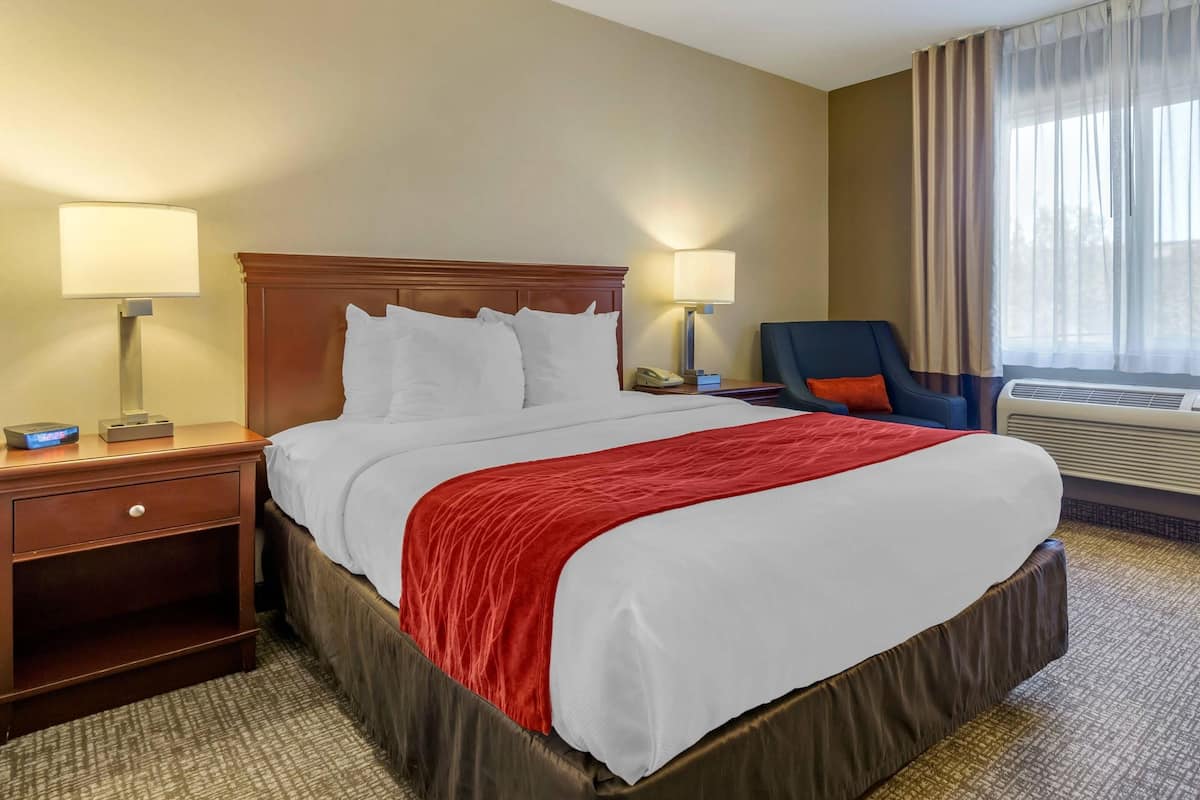 A hotel room with a large bed, two bedside lamps, a chair, a desk with a phone, and an air conditioning unit.