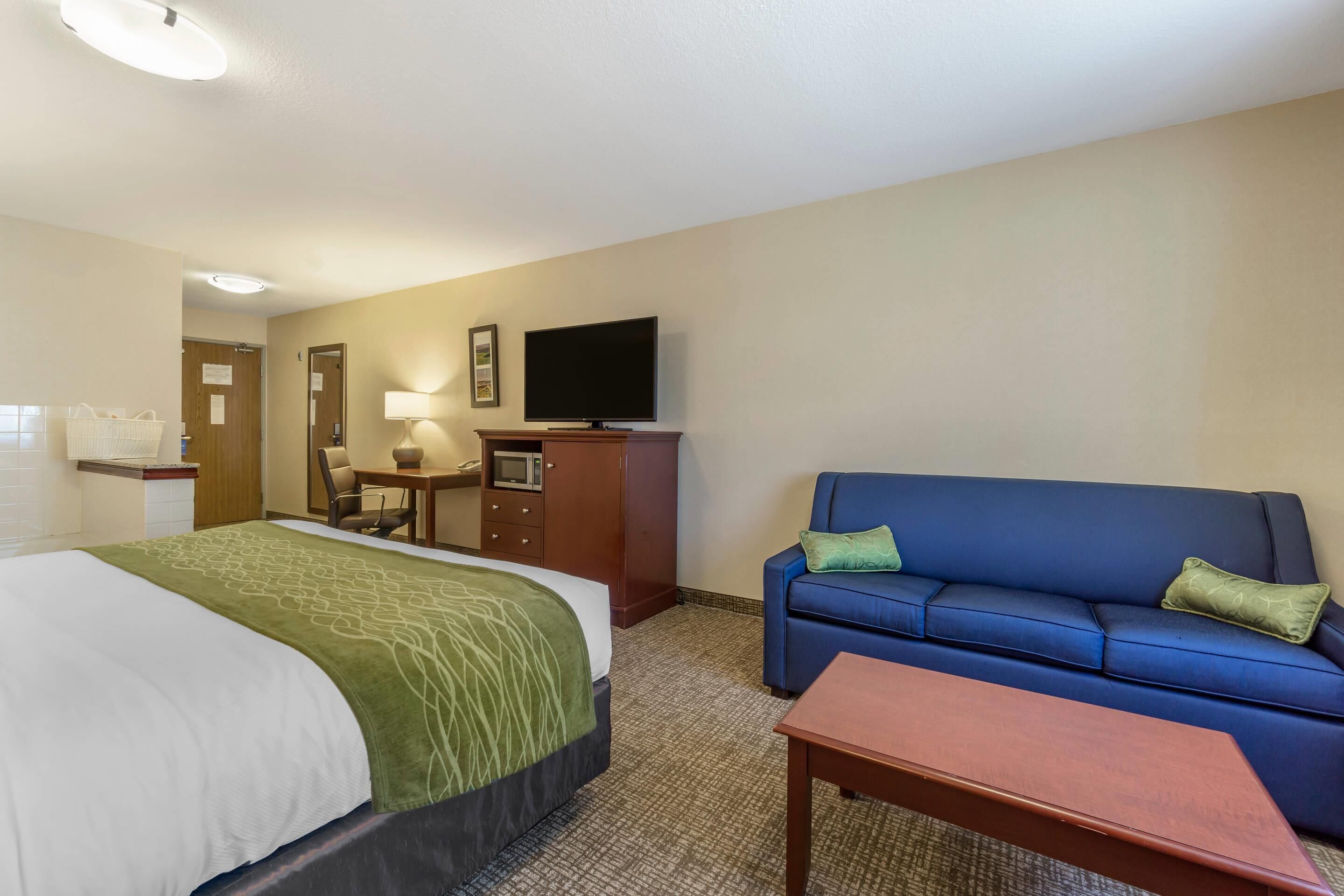 A hotel room with a bed, a blue sofa, a wooden table, a television on a cabinet, and a desk with a lamp.