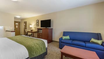 A hotel room with a bed, a blue sofa, a wooden table, a television on a cabinet, and a desk with a lamp.