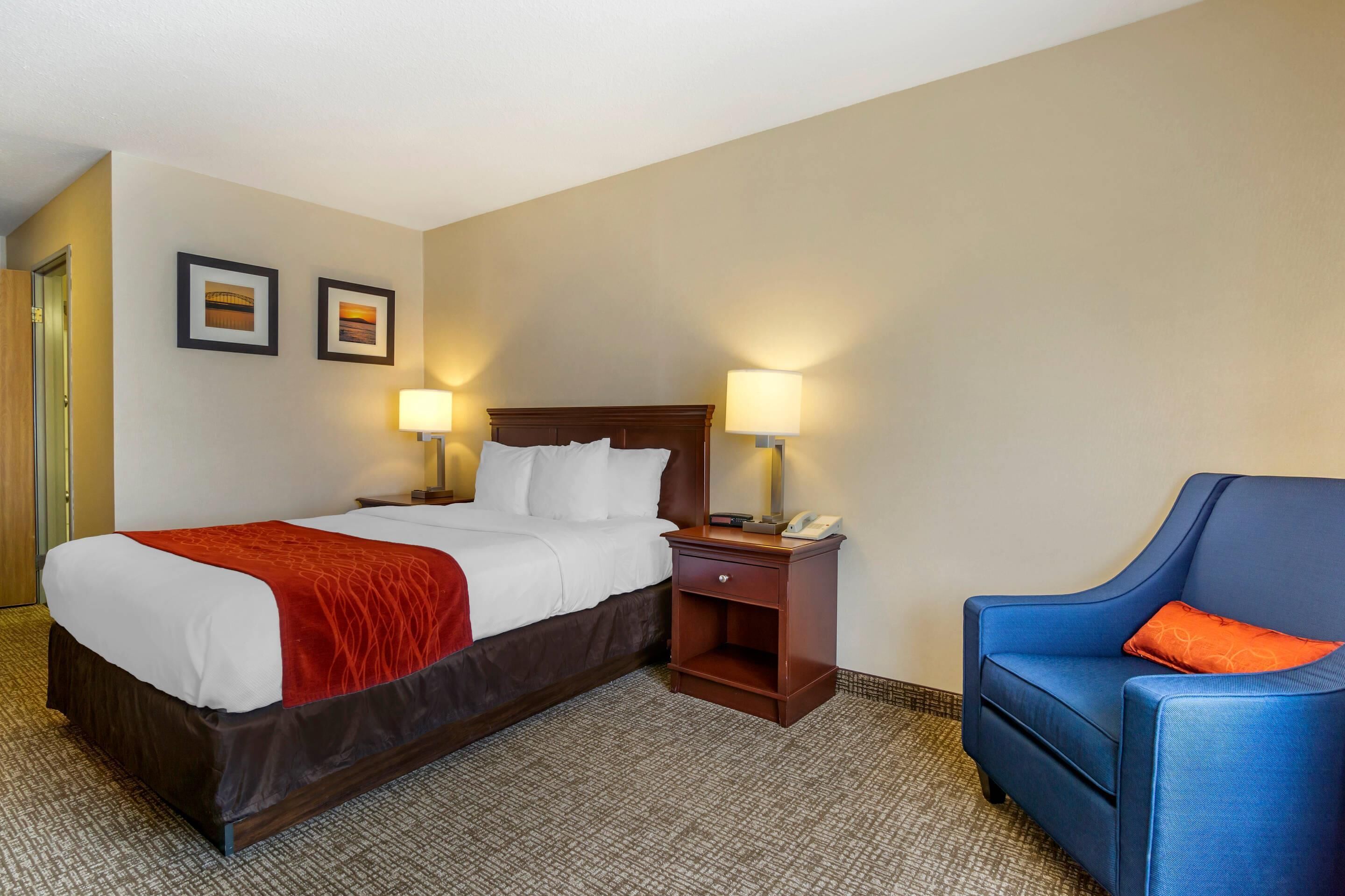 A hotel room with a large bed, two bedside lamps, a nightstand with a phone, and a blue armchair.