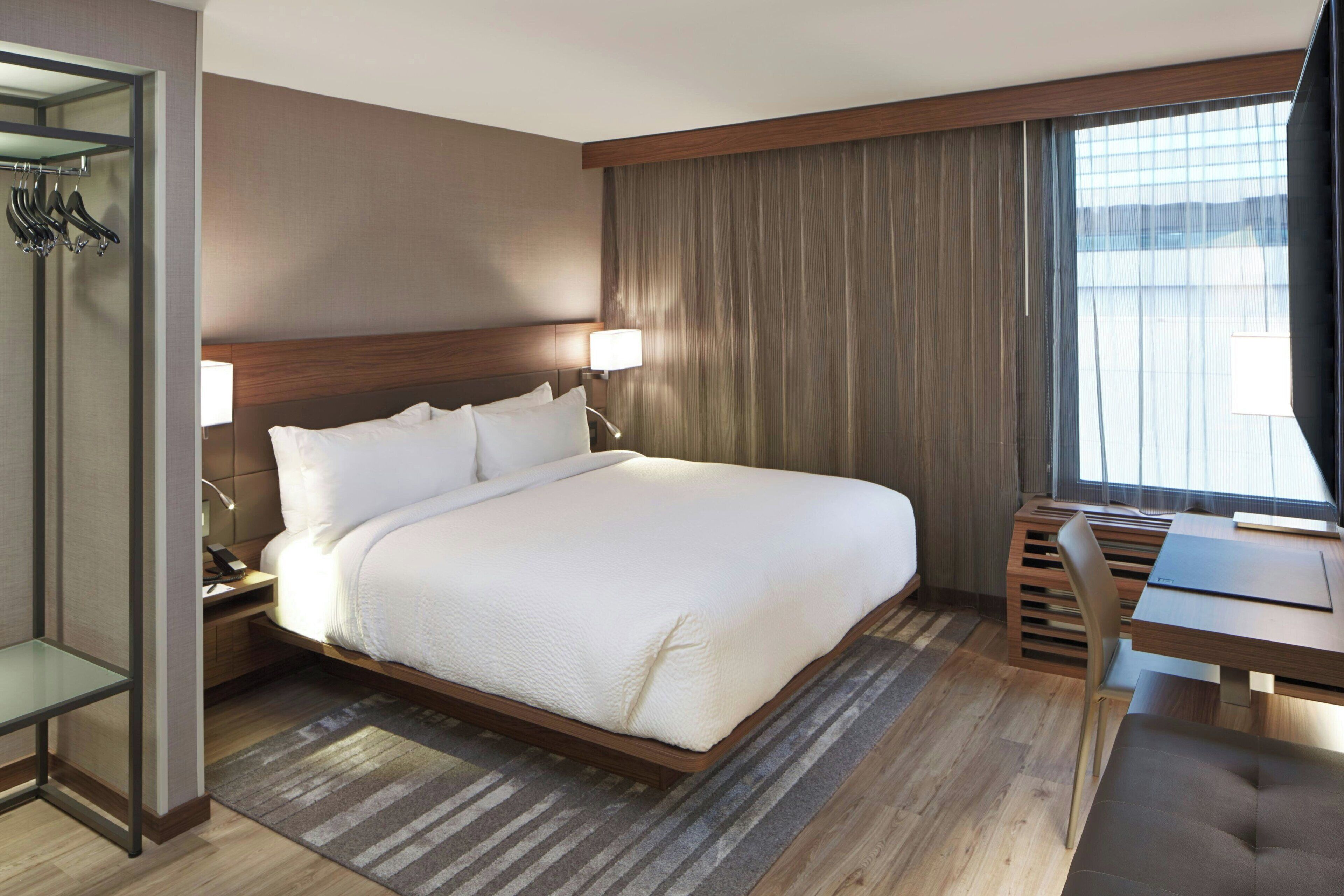 A modern hotel room with a large bed, bedside tables, a desk, and a window with curtains.
