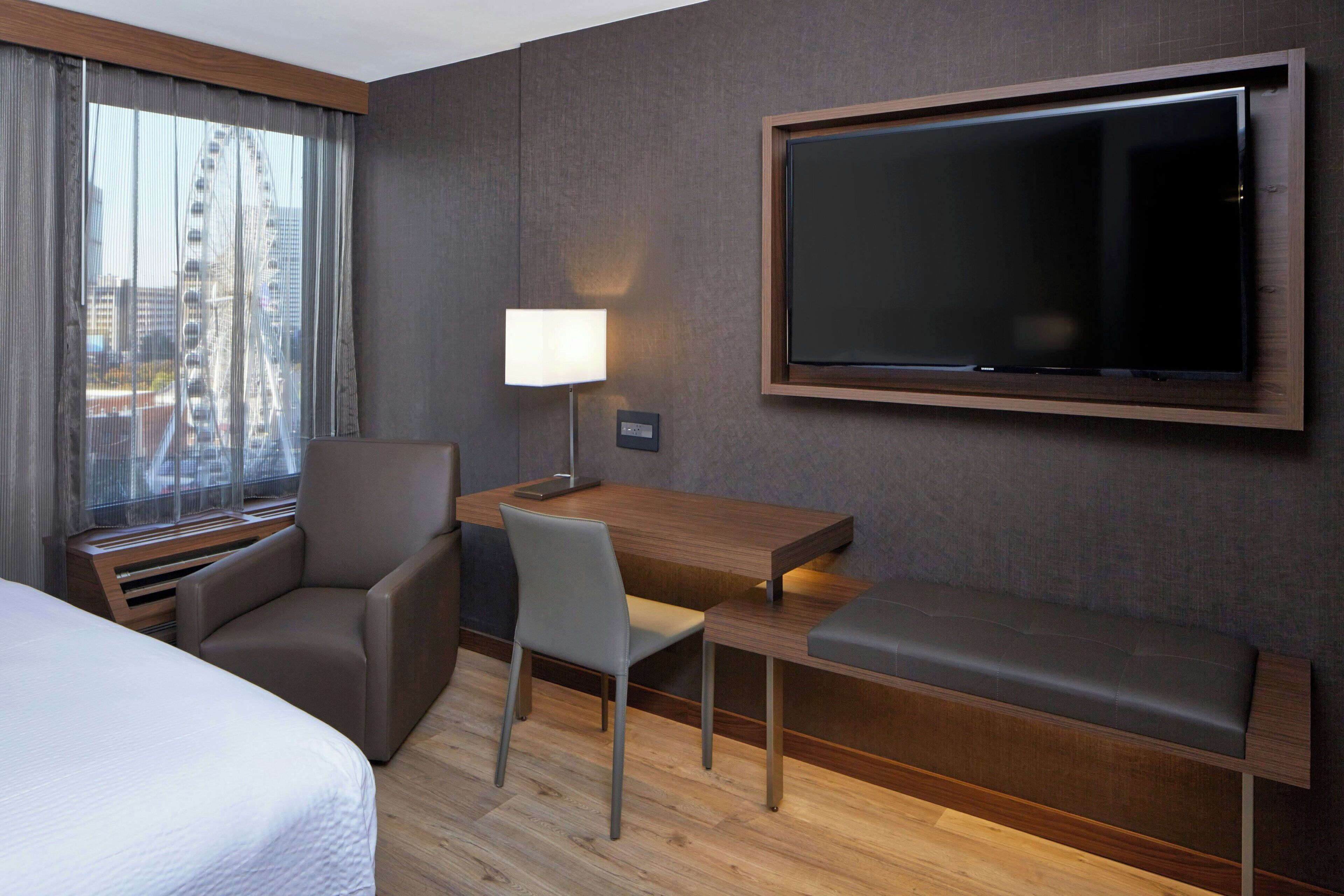 A hotel room with a bed, a desk with a chair, a TV mounted on the wall, and a window with a view of a Ferris wheel.