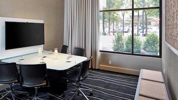 A conference room with a large flat-screen TV, a white table, four black chairs, a glass of water, and a view of a parking lot through large windows.
