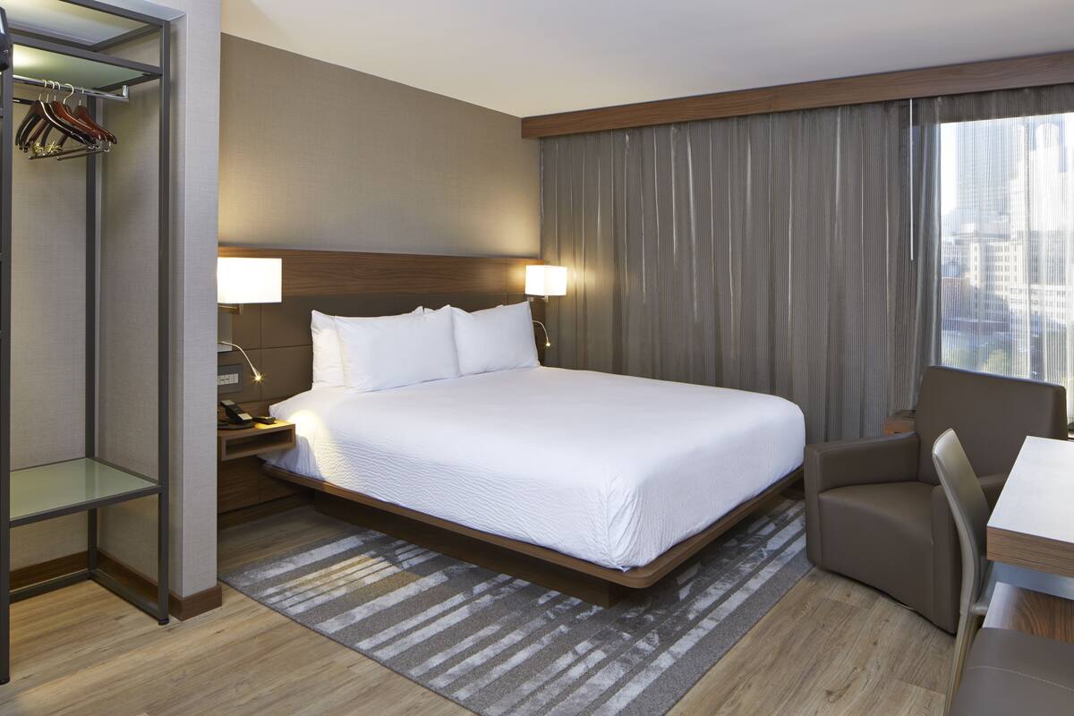 A hotel room with a large bed, a desk, a chair, a wardrobe, and a view of the city through the window.