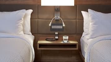 A hotel room with two beds, a bedside table with a lamp, a glass of water, and a power outlet.