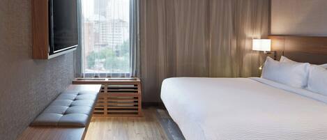 Una habitación de hotel moderna con una cama grande, un banco, un televisor montado en la pared y un ventanal con cortinas.