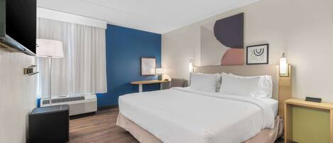 Quarto de hotel com uma cama grande, escrivaninha, cadeira, luminária e janela com cortinas.